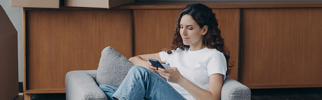 A képen egy hölgy ül egy kamapén, mobiltelefonját nézi, körülötte dobozok vannak a szobában